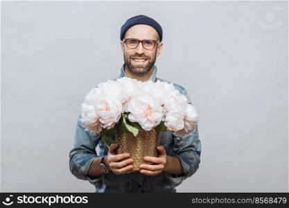 Positive unshaven male wears spectacles, holds beautiful bouquet of flowers, going to present them to his wife and congratulate with birthday, isolated over white background. People, presents concept