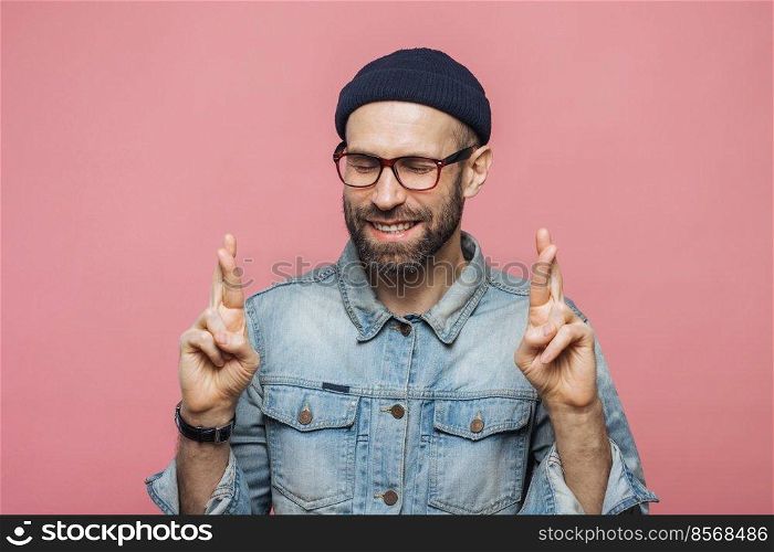 Positive unshaven male with glad look crosses fingers, keeps eyes closed, has great desire and hope for better, wears fashionable clothes, isolated over pink background. People and desire concept