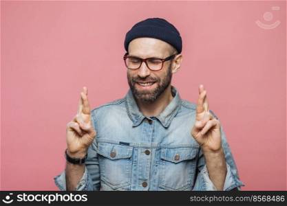 Positive unshaven male with glad look crosses fingers, keeps eyes closed, has great desire and hope for better, wears fashionable clothes, isolated over pink background. People and desire concept