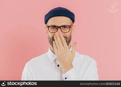 Positive unshaven man covers mouth with palm, giggles positively, wears spectacles, hears funny joke, models against pink background. Happy male designer recieves good news. Emotions concept