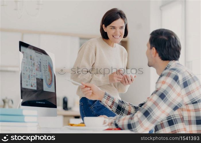 Positive woman and man discuss payment of utility bills during coffee break, man points into screen of computer, shows some graphics and diagrams, work at home in coworking space. Collaboration