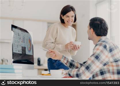 Positive woman and man discuss payment of utility bills during coffee break, man points into screen of computer, shows some graphics and diagrams, work at home in coworking space. Collaboration
