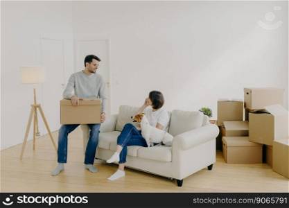 Positive woman and man pose in empty spacious room during relocation day, husband carries cardboard boxes with belongings, wife has telephone conversation, sits on comfortable sofa with dog.