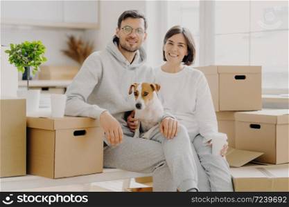Positive woman and man pose near stack of cardboard boxes, pose for making portrait with dog, relocate in new living place, wear white casual clothes, pose in spacious kitchen with big windows