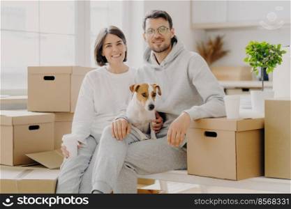 Positive woman and man pose near stack of cardboard boxes, pose for making portrait with dog, relocate in new living place, wear white casual clothes, pose in spacious kitchen with big windows