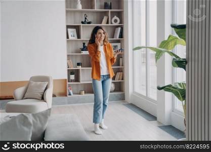 Positive woman answering call, enjoying business conversation, holding smartphone standing in modern apartment. Smiling female businessperson wearing earphones communicating by phone at home.. Smiling woman answering call, enjoying business conversation by phone in modern apartment at home