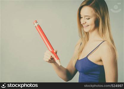 Positive woman blonde student girl holding big red pencil pen drawing. Studio shot on grey, filtered photo. Positive woman holds big pencil in hand