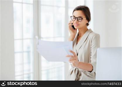 Positive woman in formal clothes, calls to business partner, discusses financial startup project, reads information from papers, wears optical glasses, stands in office, works at international company