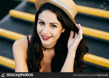 Positive woman with dark hair, shining eyes and red lips wearing straw hat and necklace feeling relaxation and having good mood while sitting at stairs. People, lifestyle, youth, fashion concept