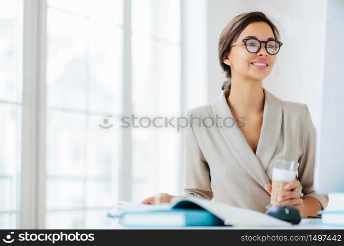 Positive woman works in office, sits at desktop, concentrated into monitor, drinks fresh milkshake, smiles and enjoys working, poses against office interior thinks about organization of business event