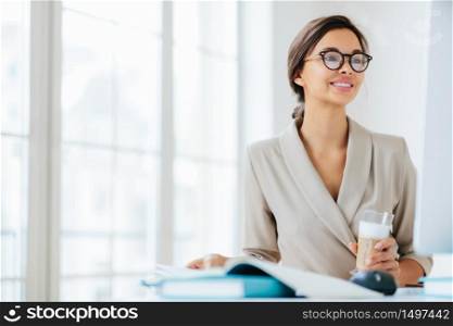 Positive woman works in office, sits at desktop, concentrated into monitor, drinks fresh milkshake, smiles and enjoys working, poses against office interior thinks about organization of business event