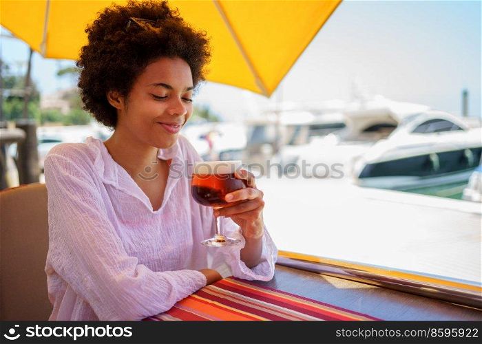 Positive young African American woman in white blouse with curly hair smiling and looking at foamy beer in glass, while sitting at table of street restaurant on sunny summer day on embankment.. Young black female drinking beer in street restaurant