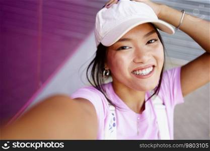Positive young Asian woman in stylish clothes touching head and looking at camera with smile while taking selfie on city street in daytime. Glad Asian female taking selfie