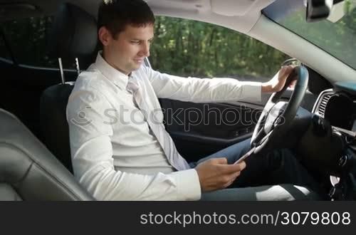 Positive young business executive browsing the net on mobile phone while sitting in luxury car. Side view. Smiling businessman texting on smartphone while traveling to work by auto. Slow motion. Steadicam stabilized shot.