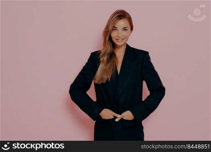 Positive young business woman keeping hands in pockets of black formal suit, being satisfied with working day, posing with magnificent smile on face against pink background, demonstrating confidence. Happy businesswoman smiling at camera on pink background