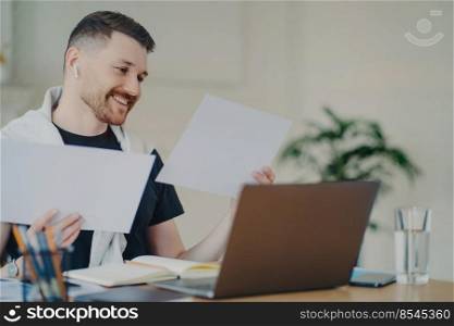 Positive young businessman in casual wear analyzing project results and feeling happy, using modern laptop at his cozy workplace at home. Successful entrepreneur feeling satisfied with business result. Happy entrepreneur looking at documents while working at home office