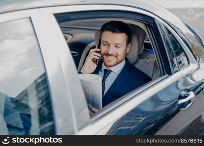 Positive young company executive reading latest newspaper, sharing good news regarding with colleague or partner, satisfied with publicity while riding in back of car with open window. Happy young business owner reading newspaper in car
