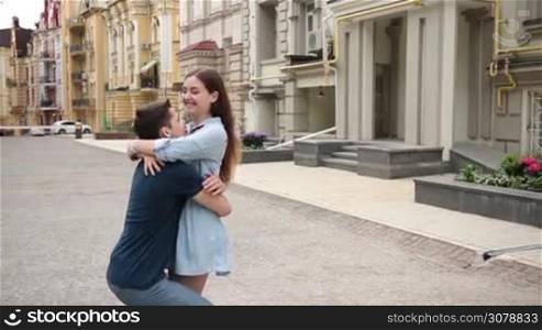 Positive young couple in love spinning around during romantic date in city street. Happy young woman embracing boyfriend&acute;s neck delicately while man holding her waist tightly in hands and whirling beloved girl. Slow motion. Stabilized shot.
