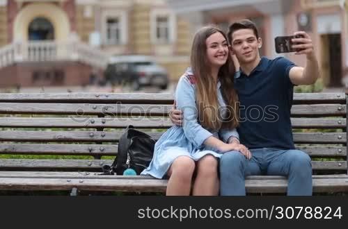 Positive young couple in love taking selfie on mobile phone while sitting on the bench in park during romantic date. Joyful hipster couple making self portrait on smarthone while relaxing together outdoors on sunny day.
