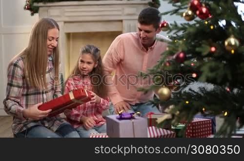 Positive young family of three exchanging gifts near Christmas tree while spending winter holidays at home. Joyful parents and excited adorable daughter sitting near xmas tree and exchanging different presents in festive decorated room. Dolly shot.
