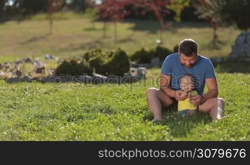 Positive young father and his smiling toddler son sitting barefoot on green grassy lawn, clapping hands and having fun over amazing summer landscape background. Joyful father and adorable infant baby boy enjoying leisure together in public park.