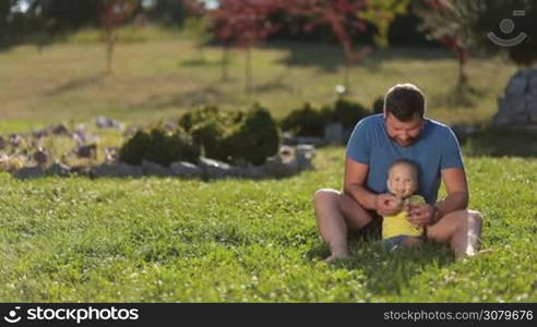 Positive young father and his smiling toddler son sitting barefoot on green grassy lawn, clapping hands and having fun over amazing summer landscape background. Joyful father and adorable infant baby boy enjoying leisure together in public park.