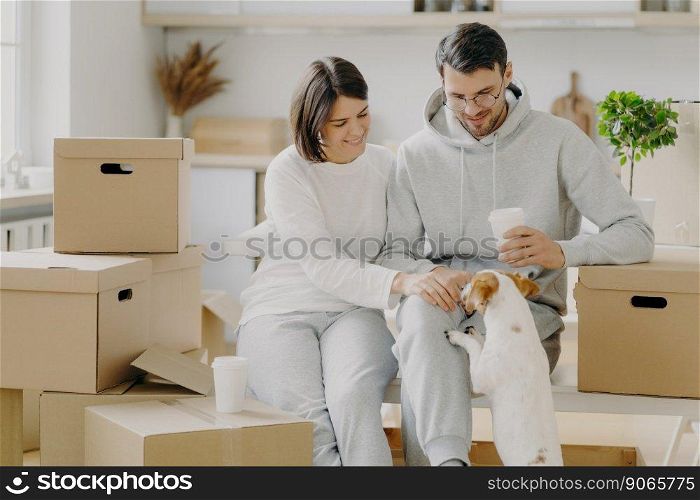 Positive young husband and wife play with dog, sit on cardboard boxes, drink takeaway coffee, have break during moving day and unpacking things, wear casual outfit, enjoy living in new flat.