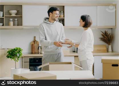 Positive young husband and wife unpack personal belongings in kitchen, carry plates, look gladfully at each other, poses indoor against cozy interior, happy to move into new modern flat or house