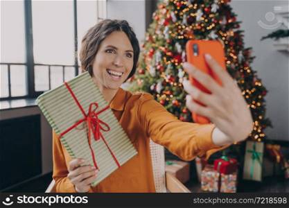 Positive young Italian woman online blogger making selfie with handmade gift on background of Xmas tree decorated with colored lights and balls while relaxing at home. People and christmas holidays. Young Italian woman online blogger making selfie with christmas gift on background of Xmas tree