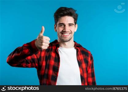 Positive young man smiles to camera. Hipster guy showing thumb up sign over blue background. Winner. Success. Body language. Positive young man smiles to camera. Hipster guy showing thumb up sign over blue