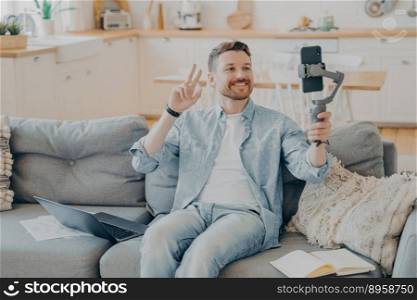 Positive young man with beard showing peace sign during video call, sitting on comfy couch with laptop and papers, open note book next to him, blurred kitchen counter in background. Positive young man showing peace sign during video call while sitting on couch
