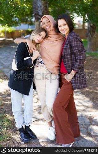 Positive young multiethnic female best friends in casual clothes smiling and looking at camera while cuddling in park on sunny day. Delighted young diverse ladies embracing and smiling in park