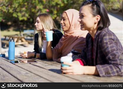 Positive young multiethnic female friends in casual clothes smiling while resting in outdoor cafe and drinking coffee from disposable cups. Cheerful diverse ladies having coffee break and chatting in city park