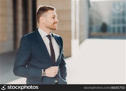 Positive young prosperous unshaven male manager wears formal clothes, has walk outdoor, concentrated into distance, hurries for business meeting with his colleagues. People and career concept