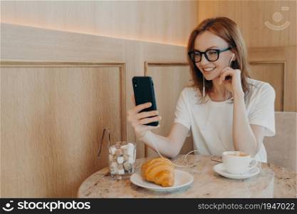 Positive young redhead female blogger has video chat with followers holds modern mobile phone and earphones sits at table in cafeteria drinks coffee eats croissant uses free internet connection. Positive young redhead female blogger has video chat with followers holds modern mobile phone