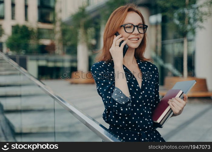 Positive young redhead woman enjoys mobile phoning keeps smartphone near ear looks into distance holds laptop computer and diary wears eyeglasses polka dot dress waits for meeting in street.. Positive young redhead woman enjoys mobile phoning keeps smartphone near ear