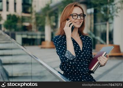 Positive young redhead woman enjoys mobile phoning keeps smartphone near ear looks into distance holds laptop computer and diary wears eyeglasses polka dot dress waits for meeting in street.. Positive young redhead woman enjoys mobile phoning keeps smartphone near ear