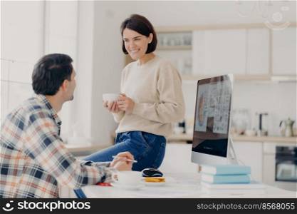 Positive young woman and man discuss renting apartment, pose in coworking space with computer and paper documents, happy wife drinks coffee, looks with pleasant smile at husband, manage budget