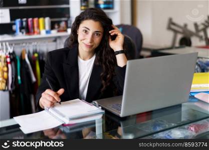 Positive young woman, in smart casual clothes, looking at camera with smile and touching dark hair, while sitting at desk and making notes during psychology session in gym. Happy sports psychologist looking at camera