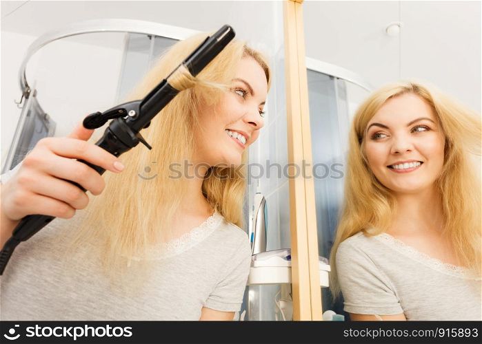 Positive young woman preparing her blonde hair, using curling pin in home bathroom. Hairdo curler creating hairstyle.. Woman using hair curler