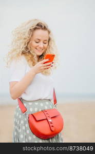 Positive young woman uses modern smart phone, has cheerful expression, makes shopping online, carries bright small bag, has outdoor walk at seaside. People, technology, positiveness concept.