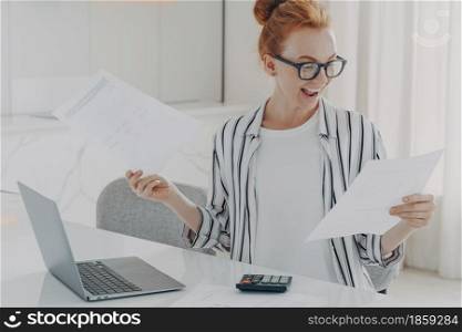 Positive young woman with natural red hair calculates utility charges engaged in planning monthly budget holds papers sits a desktop with laptop computer and calculator counts home expenses.. Positive young woman with red hair calculates utility charges engaged in planning monthly budget