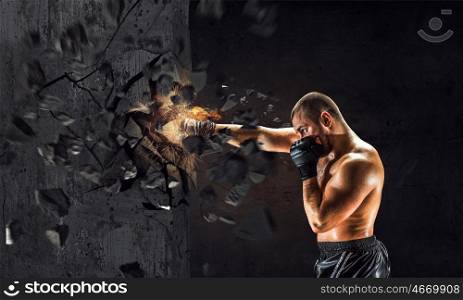 Possessing power and endurance. Strong boxer man in gloves breaking cement wall