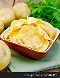 Potato chips in a clay bowl on a napkin, fresh potatoes, parsley on a wooden boards background