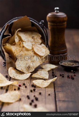Potato crisps chips snack with black pepper in steel bucket on wooden table background with mill and ground pepper.