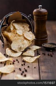 Potato crisps chips snack with black pepper in steel bucket on wooden table background with mill and ground pepper.