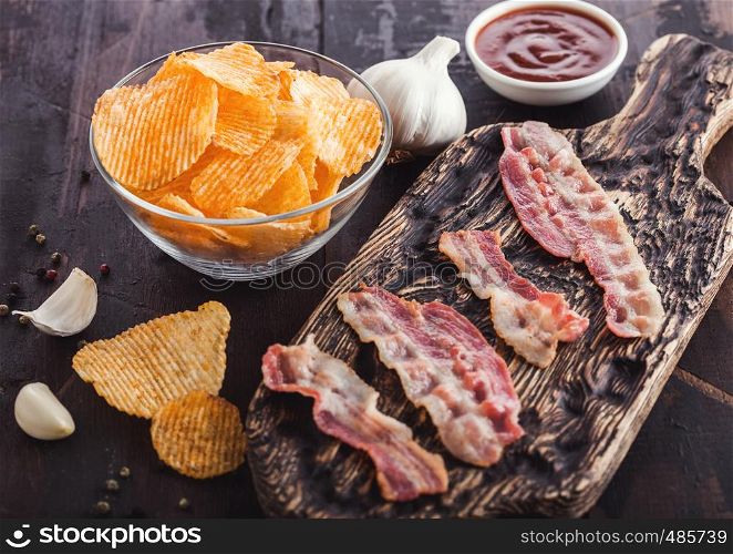 Potato crisps with backon flavour with grilled bacon rashers on vintage chopping board with garlic and sauce on wood background.