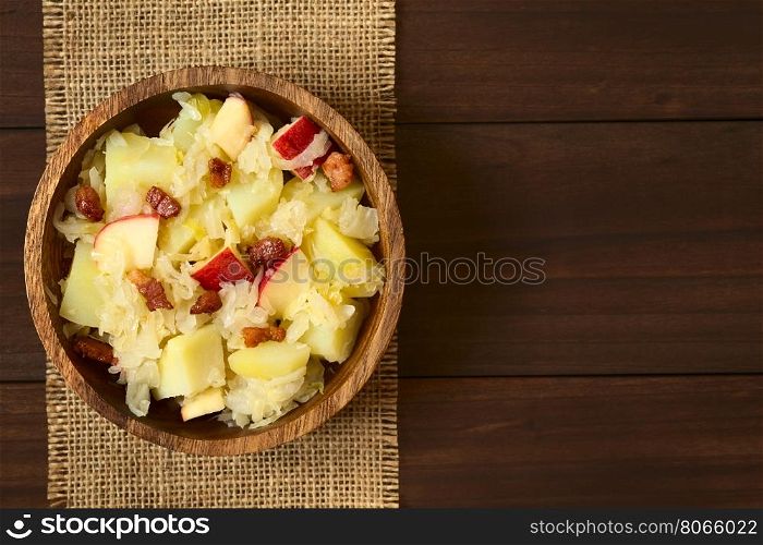 Potato, sauerkraut and apple salad with fried bacon served in wooden bowl, photographed overhead on dark wood with natural light (Selective Focus, Focus on the top of the salad)