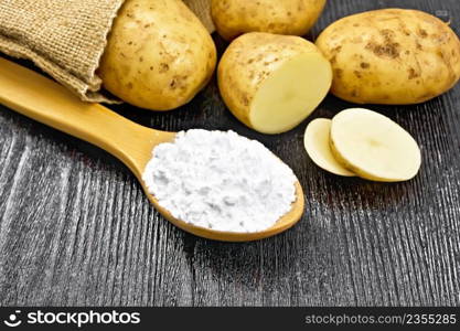 Potato starch in a spoon, vegetable tubers in a bag and on table against the background of wooden board 