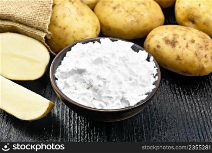 Potato starch in clay bowl, vegetable tubers in a bag and on a table against dark wooden board background